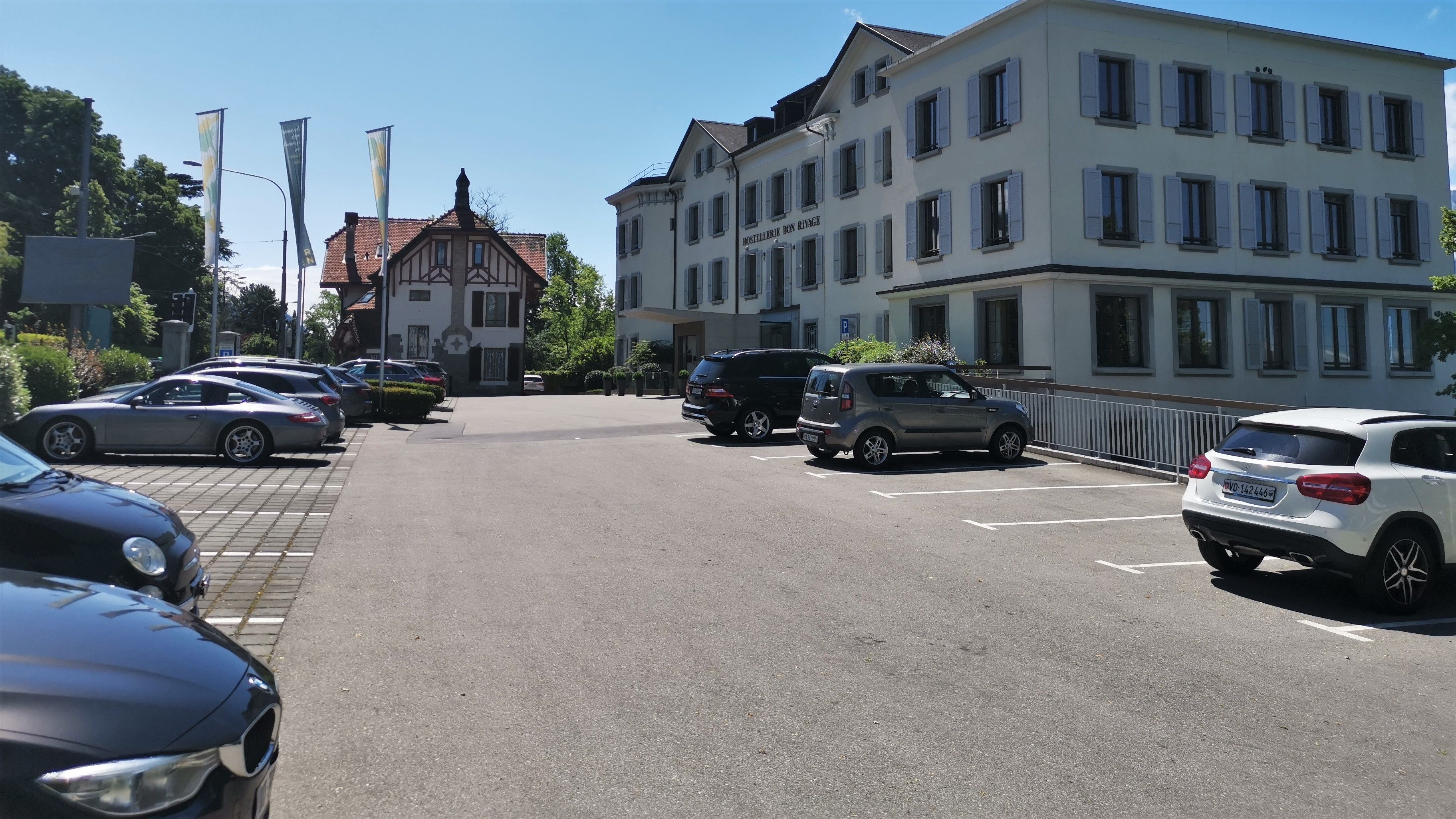 Un estacionamiento con varios coches, un edificio con un cartel que dice "HOTELIERIE DON RIVAGE" y un edificio blanco con numerosas ventanas.