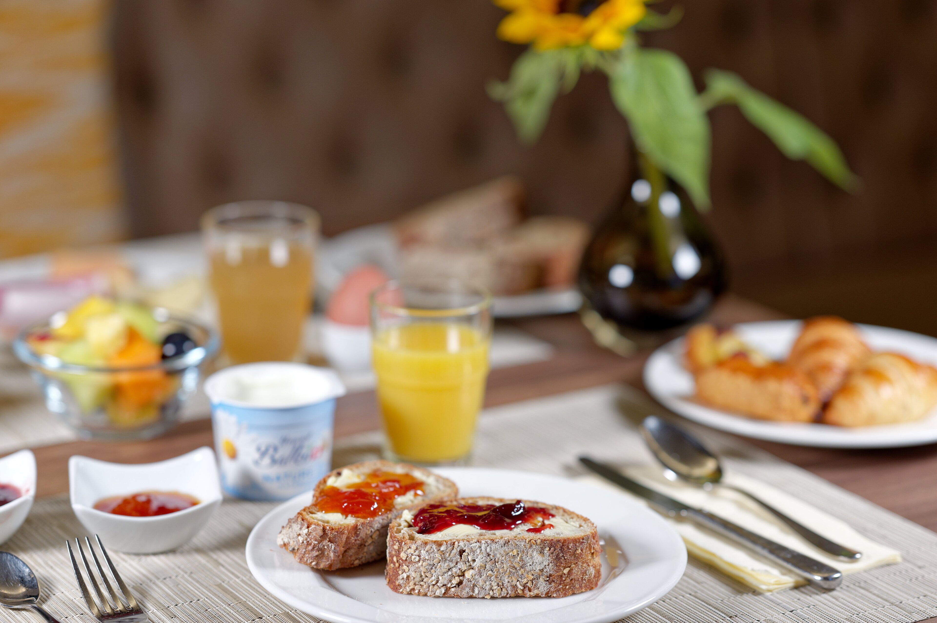 Ein Frühstückstisch mit Toast, Marmelade, Butter, einem Glas Orangensaft, einer Obstschale und einer Vase mit einer Sonnenblume.