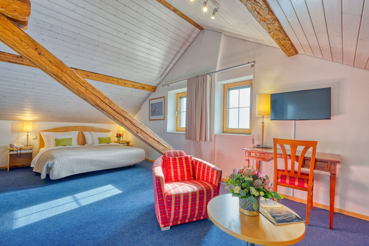 Ein Schlafzimmer mit schrägem Dach, einem Bett, einem roten Karo-Sessel, einem Schreibtisch mit Stuhl und einem an der Wand befestigten Fernseher.