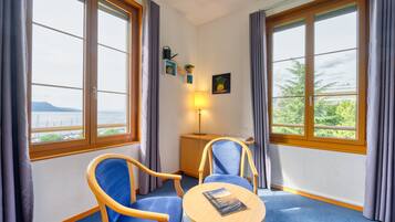 Habitación de hotel con dos sillas azules, una mesita redonda pequeña, una lámpara y vista al mar a través de ventanas grandes.