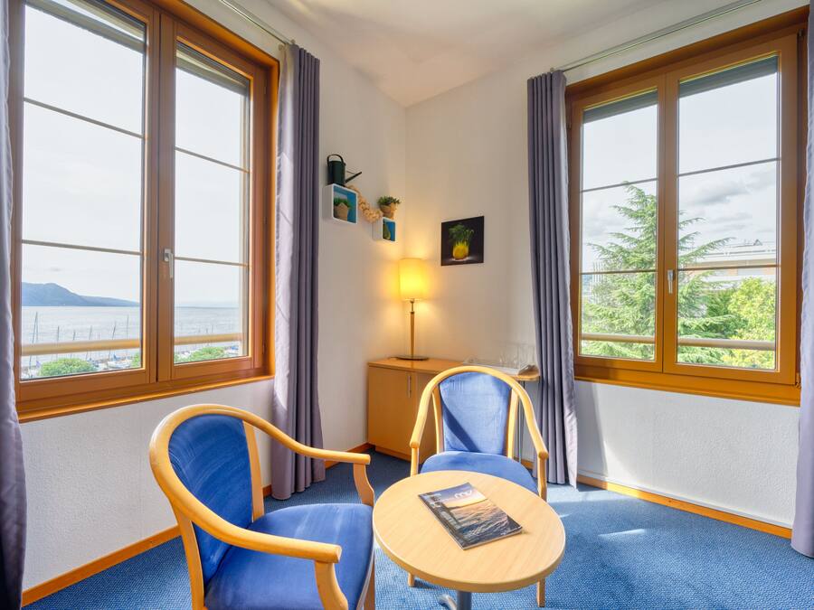 Habitación de hotel con dos sillas azules, una mesita redonda pequeña, una lámpara y vista al mar a través de ventanas grandes.