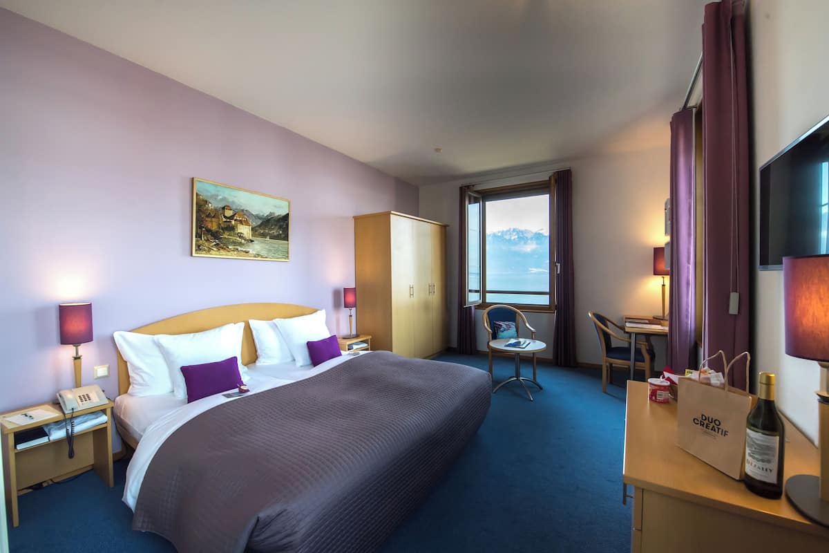 Ein Hotelzimmer mit einem großen Bett, einem Schreibtisch mit einer Flasche und einer Tasche, einem Sessel, einem Kleiderschrank und einem Bild an der Wand.