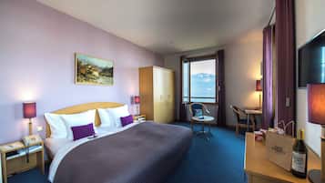 A hotel room with a large bed, a desk with a bottle and a bag, a chair, a wardrobe, and a painting on the wall.