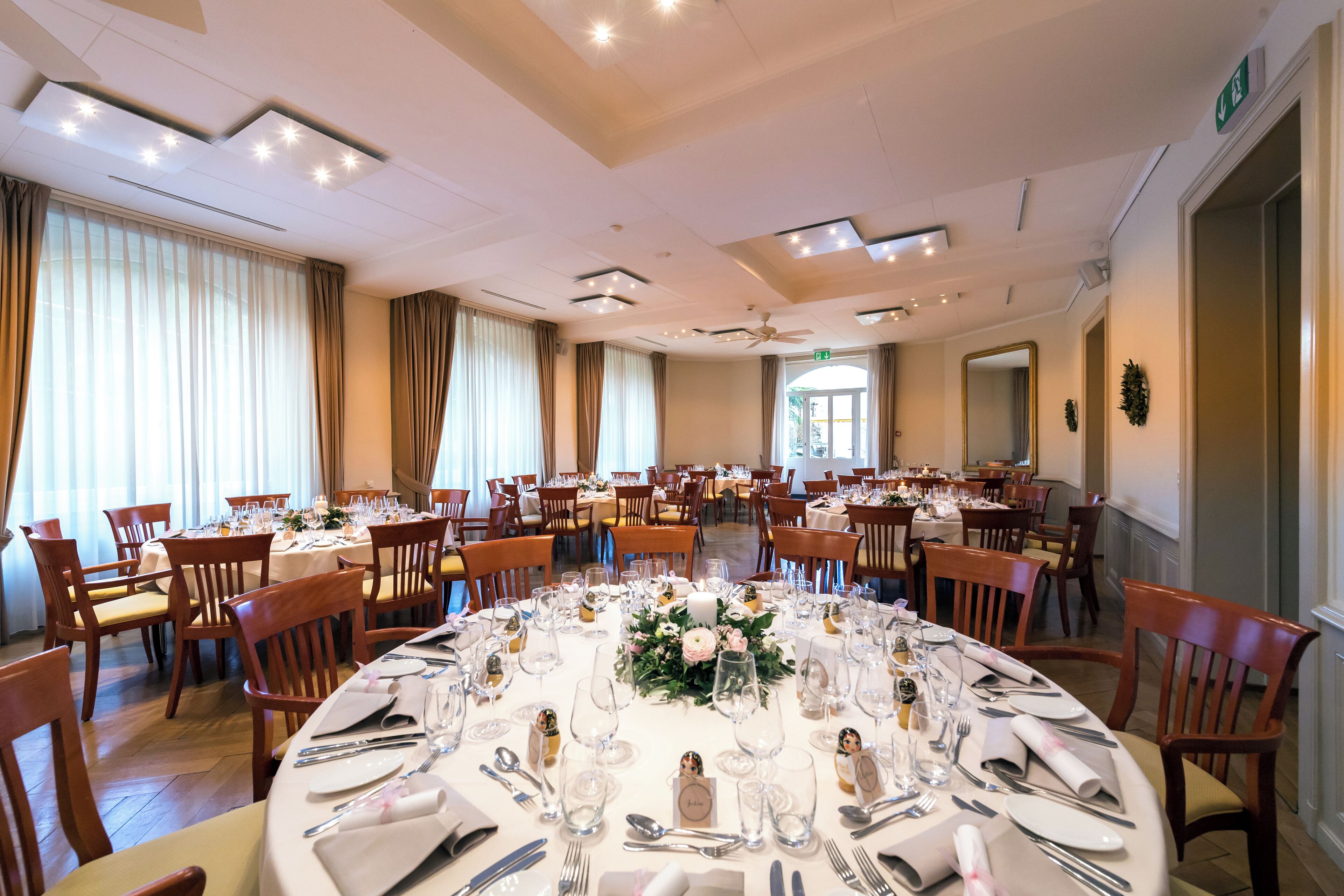 A large banquet hall with round tables set for a formal event, equipped with chandeliers and a ceiling fan.