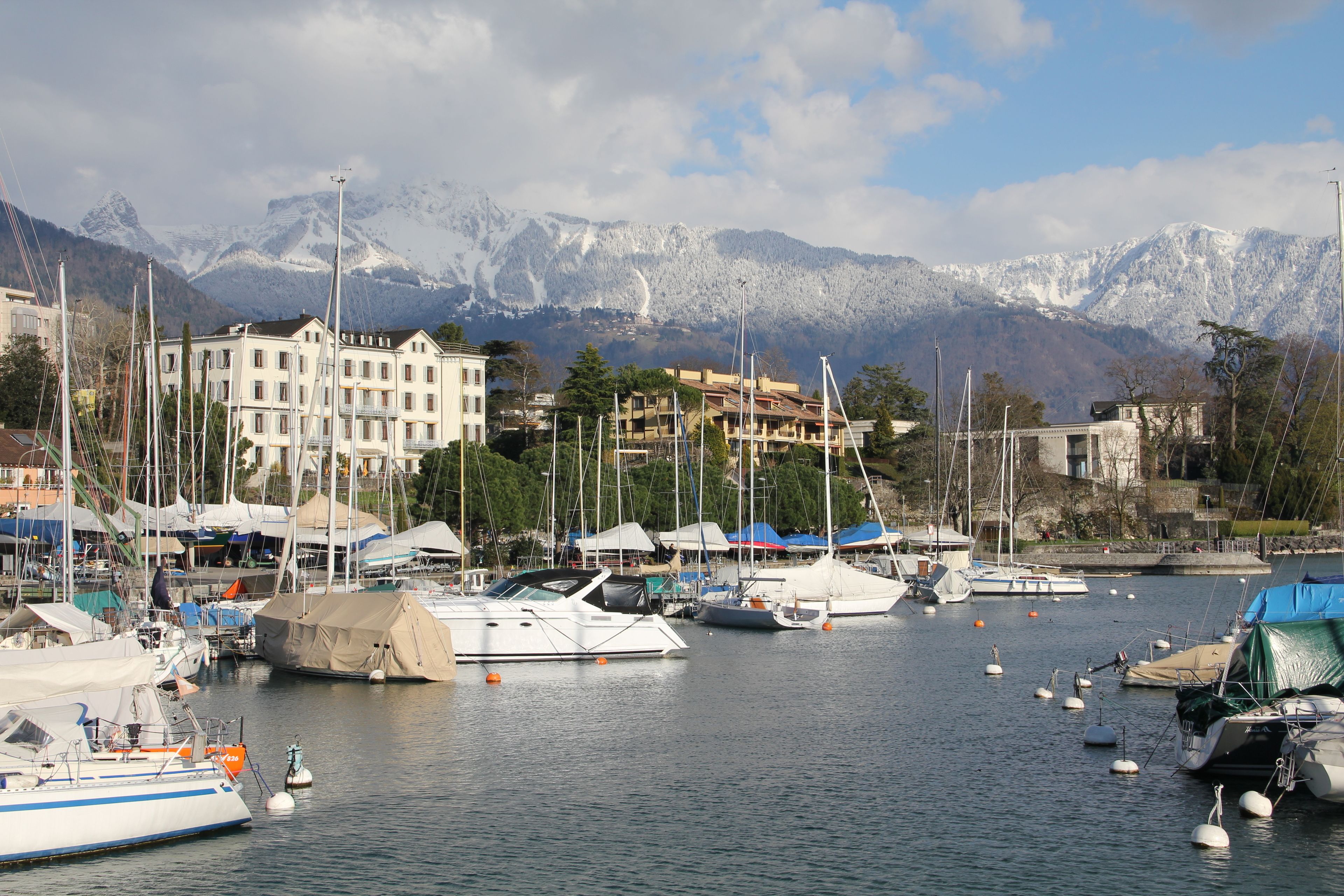 A marina with boats docked, a large building with multiple balconies, and snow-capped mountains in the background.
