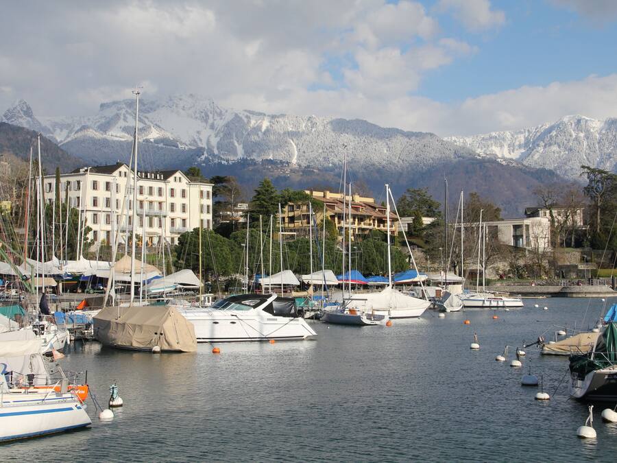 A marina with boats docked, a large building with multiple balconies, and snow-capped mountains in the background.