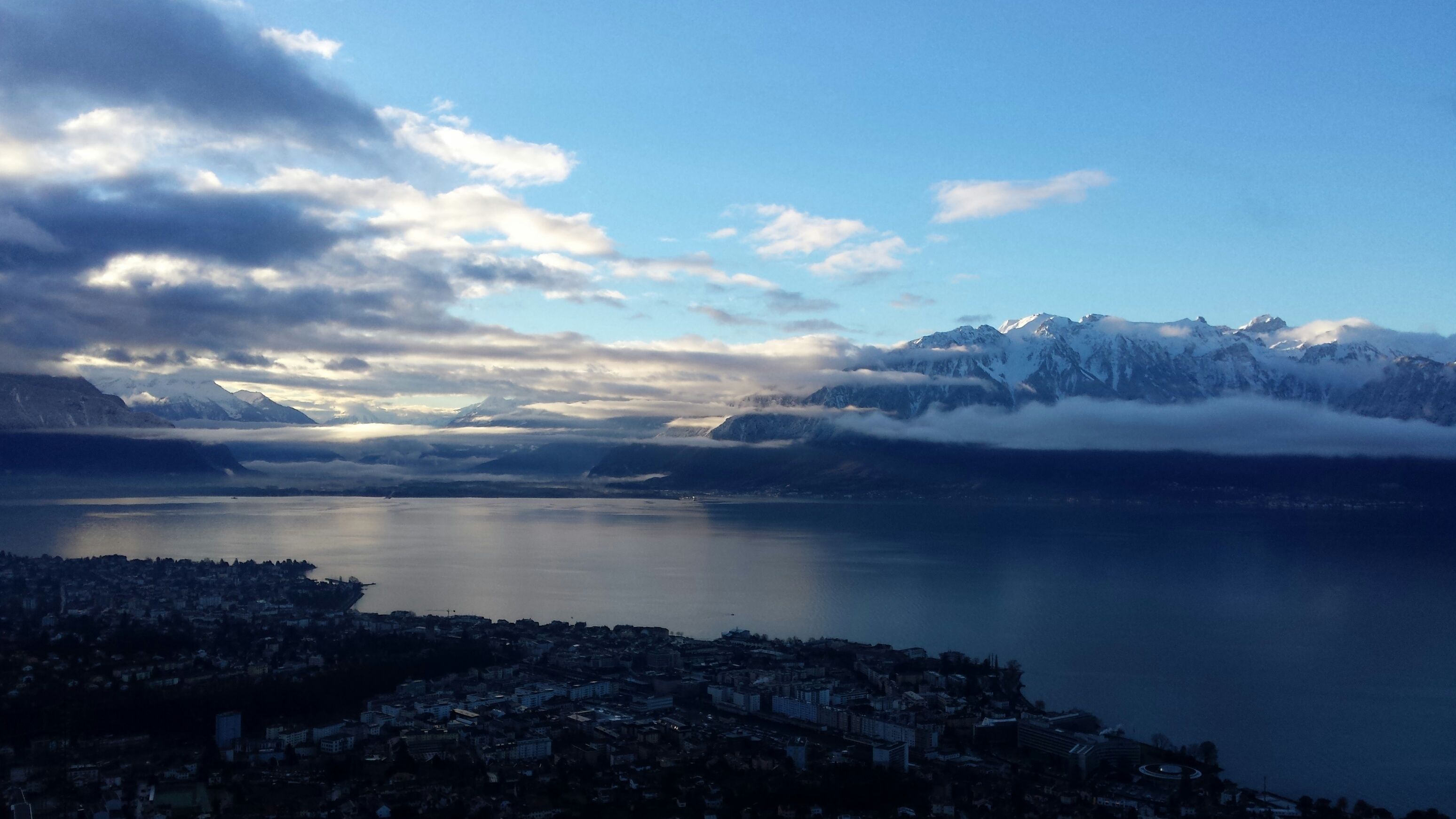Vista panorâmica de uma paisagem montanhosa com um lago e uma cidade ao primeiro plano.