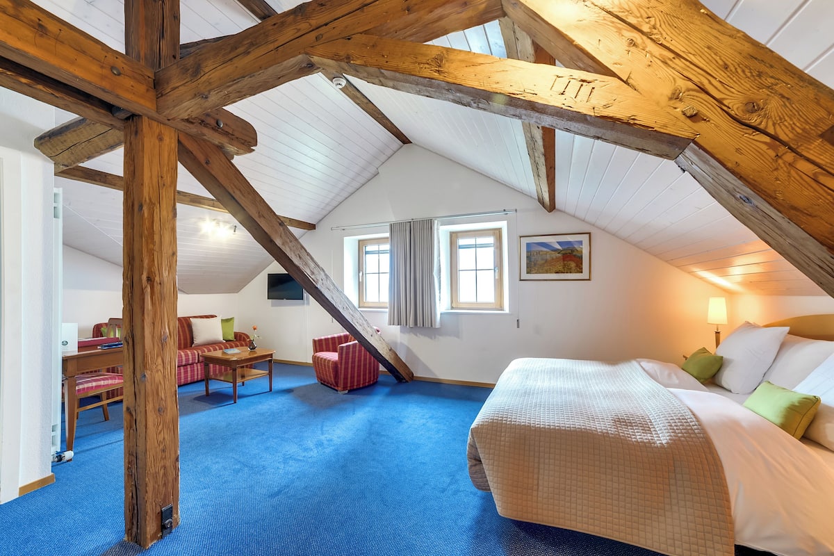 Ein Schlafzimmer mit schrägverdeckter Decke, einem Bett, einem Schreibtisch, einem Stuhl und einem Fenster.