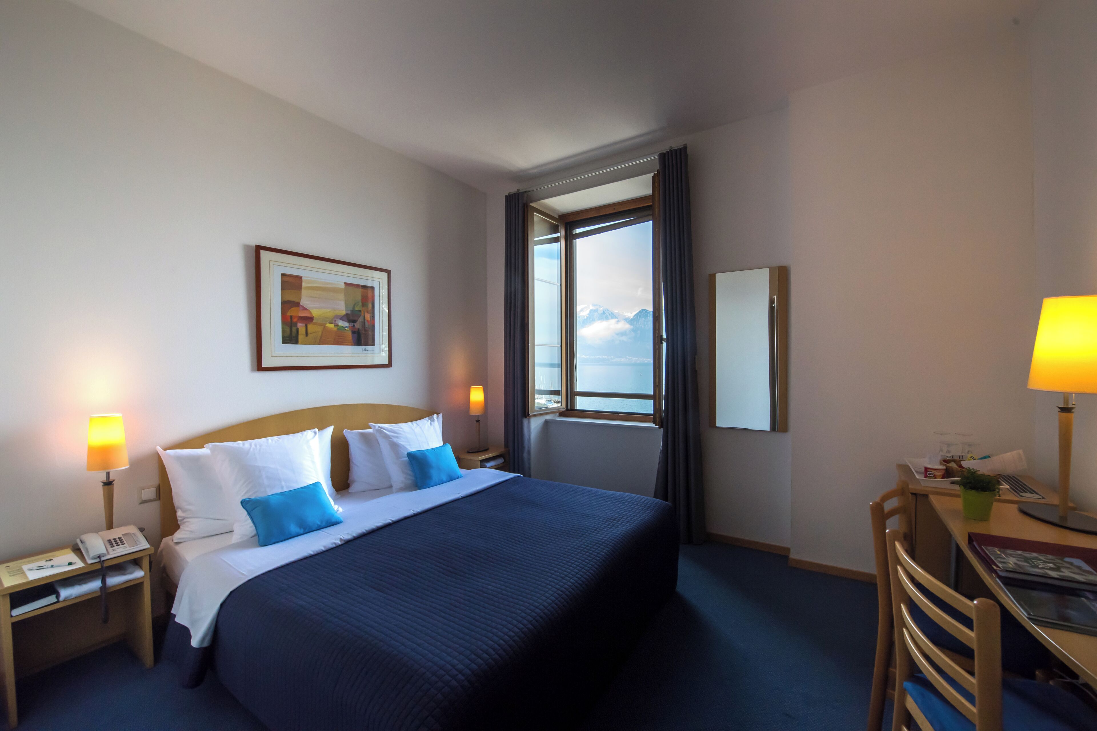 Ein Hotelzimmer mit einem großen Bett, einem Schreibtisch mit Lampe, einem Sessel, einem Telefon und einem Gemälde an der Wand.