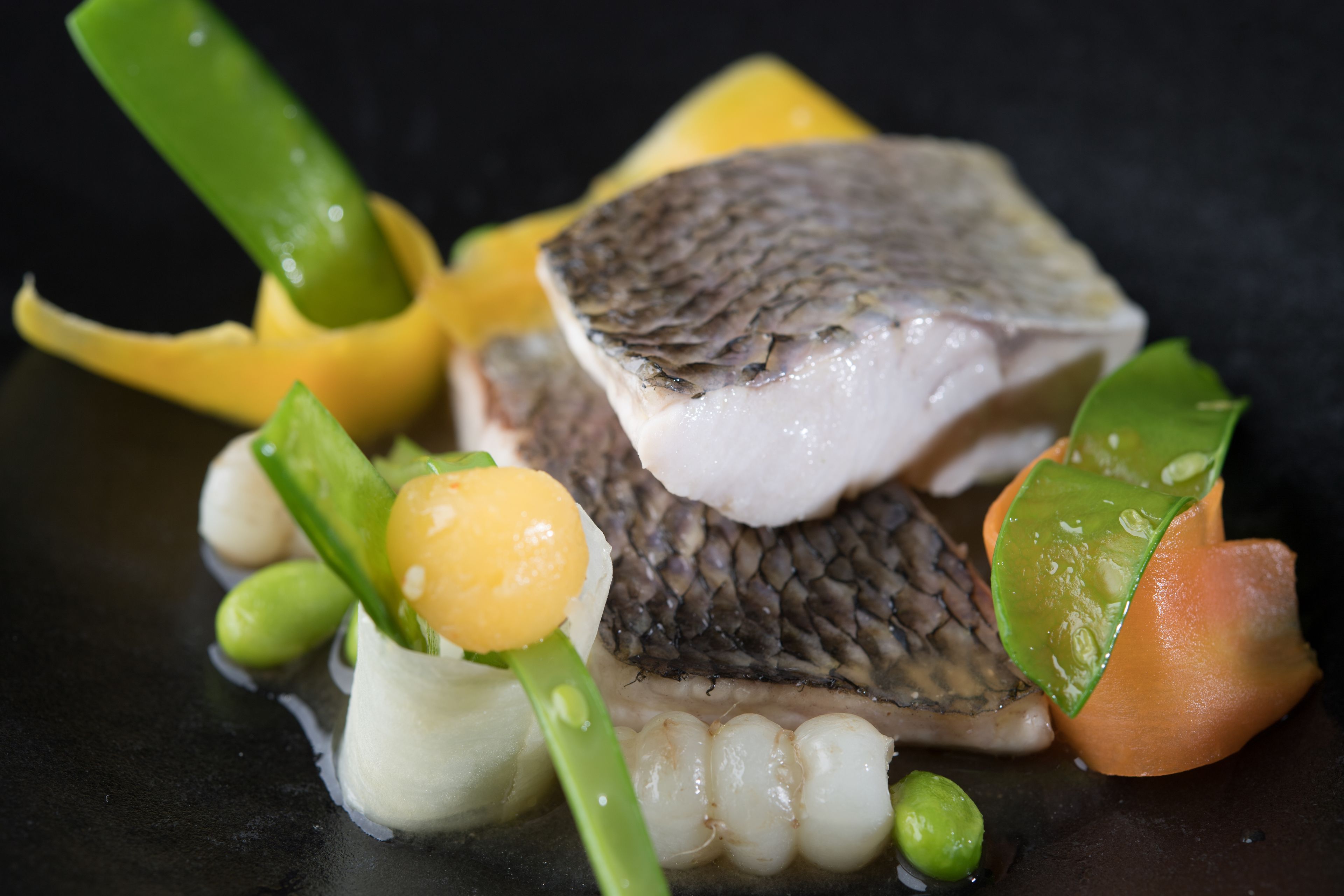 A dish with fish, vegetables, and a yellow element on a dark surface.