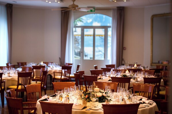 Une salle de banquet avec des tables rondes dressées pour le repas, des chaises en bois et une grande fenêtre donnant sur des palmiers.