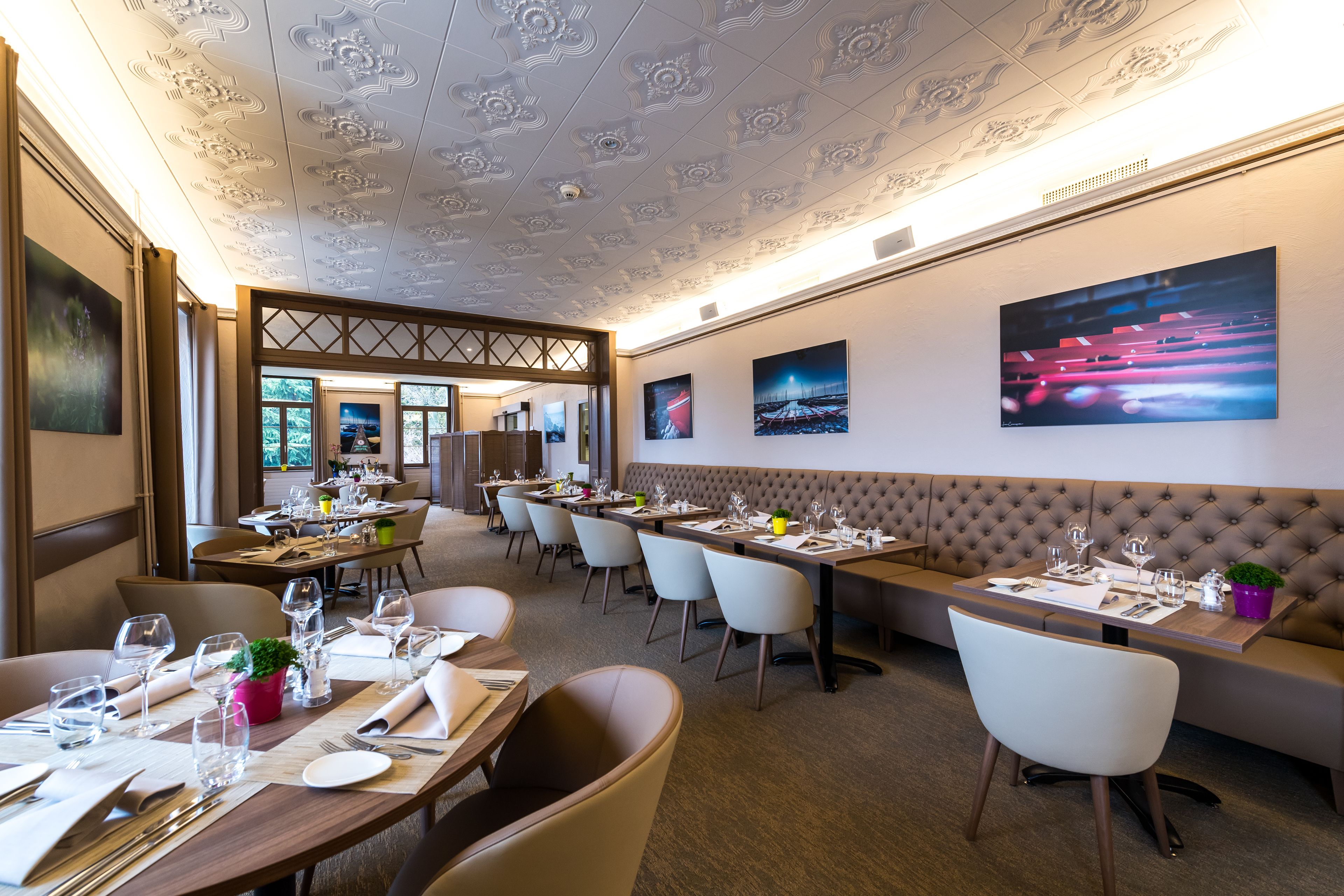 Ein Restaurant mit hoher Decke, mehreren Esstischen und Stühlen. An den Wänden befinden sich Gemälde und auf einem Tisch steht ein Blumentopf.