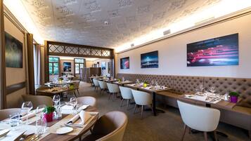 A restaurant with a high ceiling, multiple dining tables, and chairs. There are paintings on the walls and a potted plant on one table.