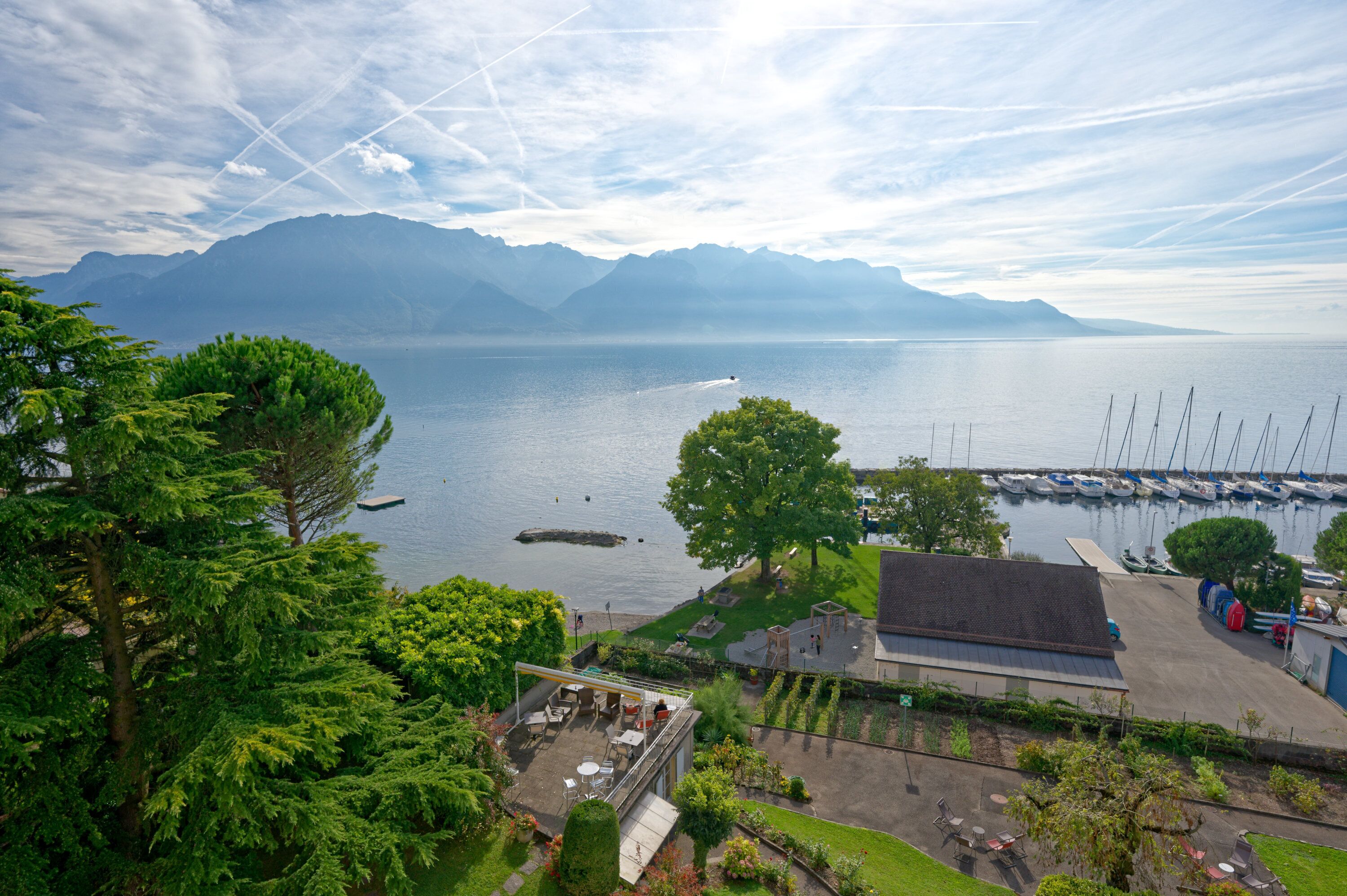 Vista de un lago con montañas al fondo, un puerto deportivo con barcos y un edificio con terraza.