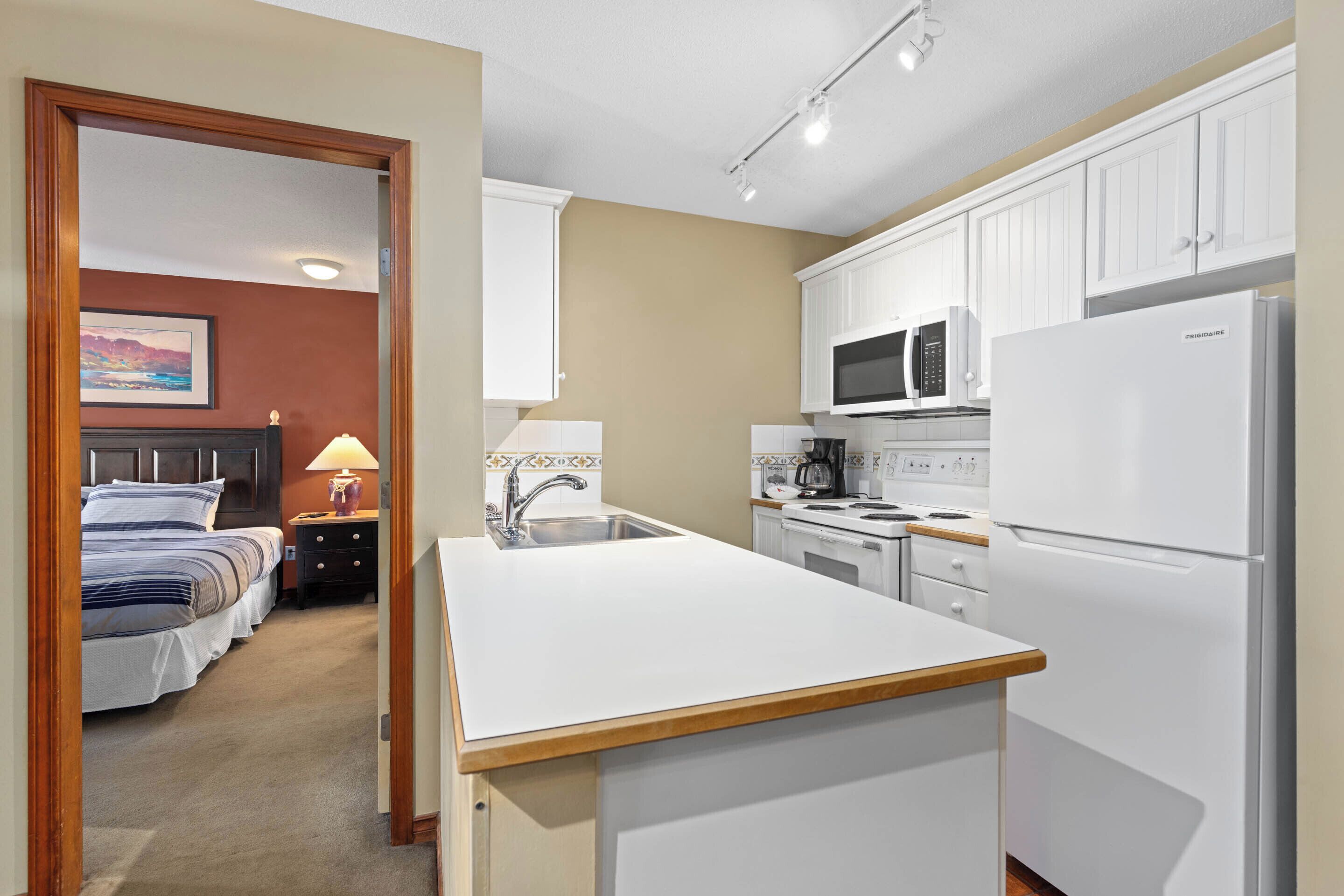 A compact hotel room with a kitchenette, a bed with a headboard, and a nightstand with a lamp.