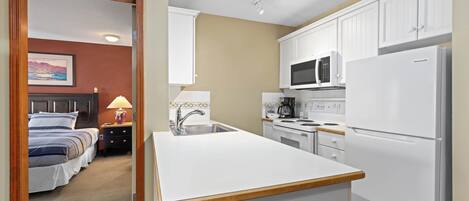 A compact hotel room with a kitchenette, a bed with a headboard, and a nightstand with a lamp.