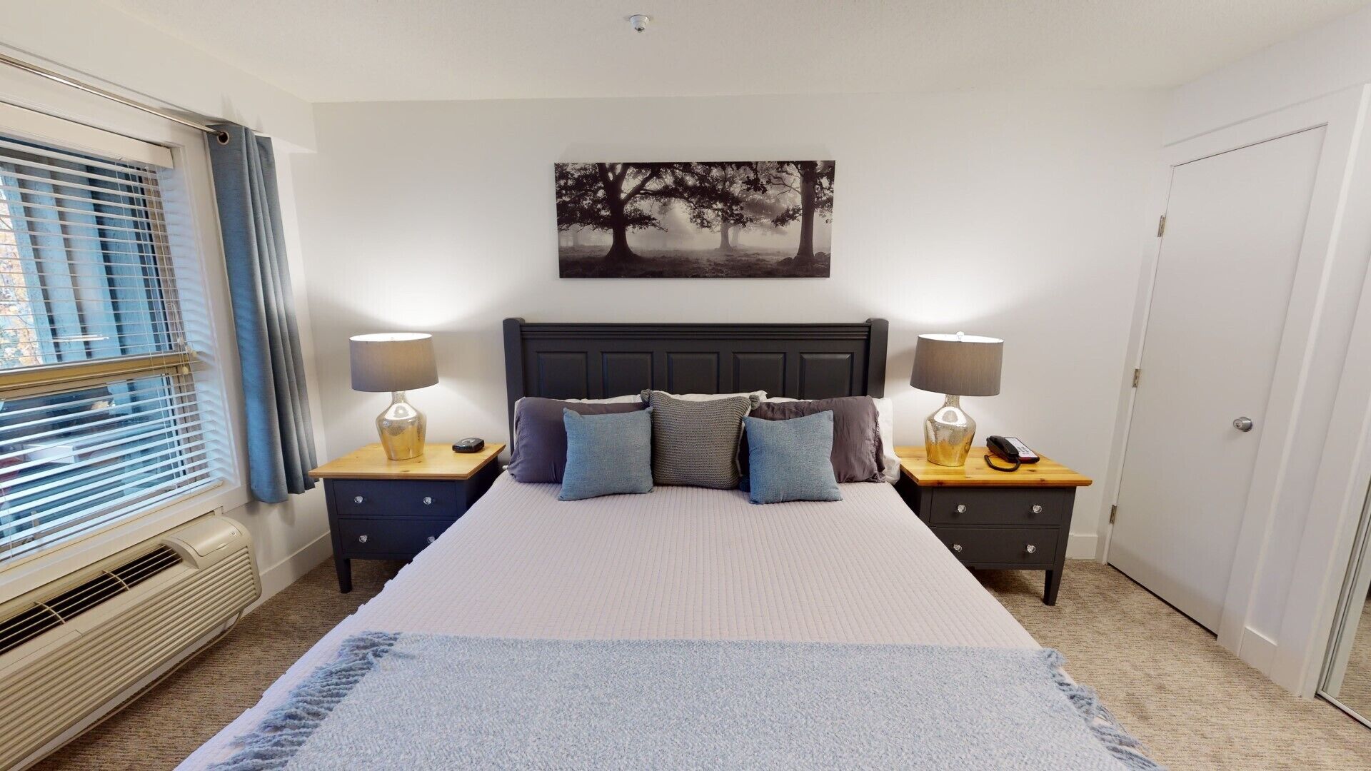 Une chambre à coucher avec un grand lit, deux tables de chevet avec des lampes, un tableau accroché au mur et une fenêtre avec des stores.