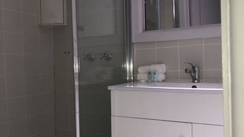 Un baño moderno con cabina de ducha de vidrio, paredes cubiertas de azulejos blancos, un tocador blanco con espejo y una ventana sobre el lavabo.