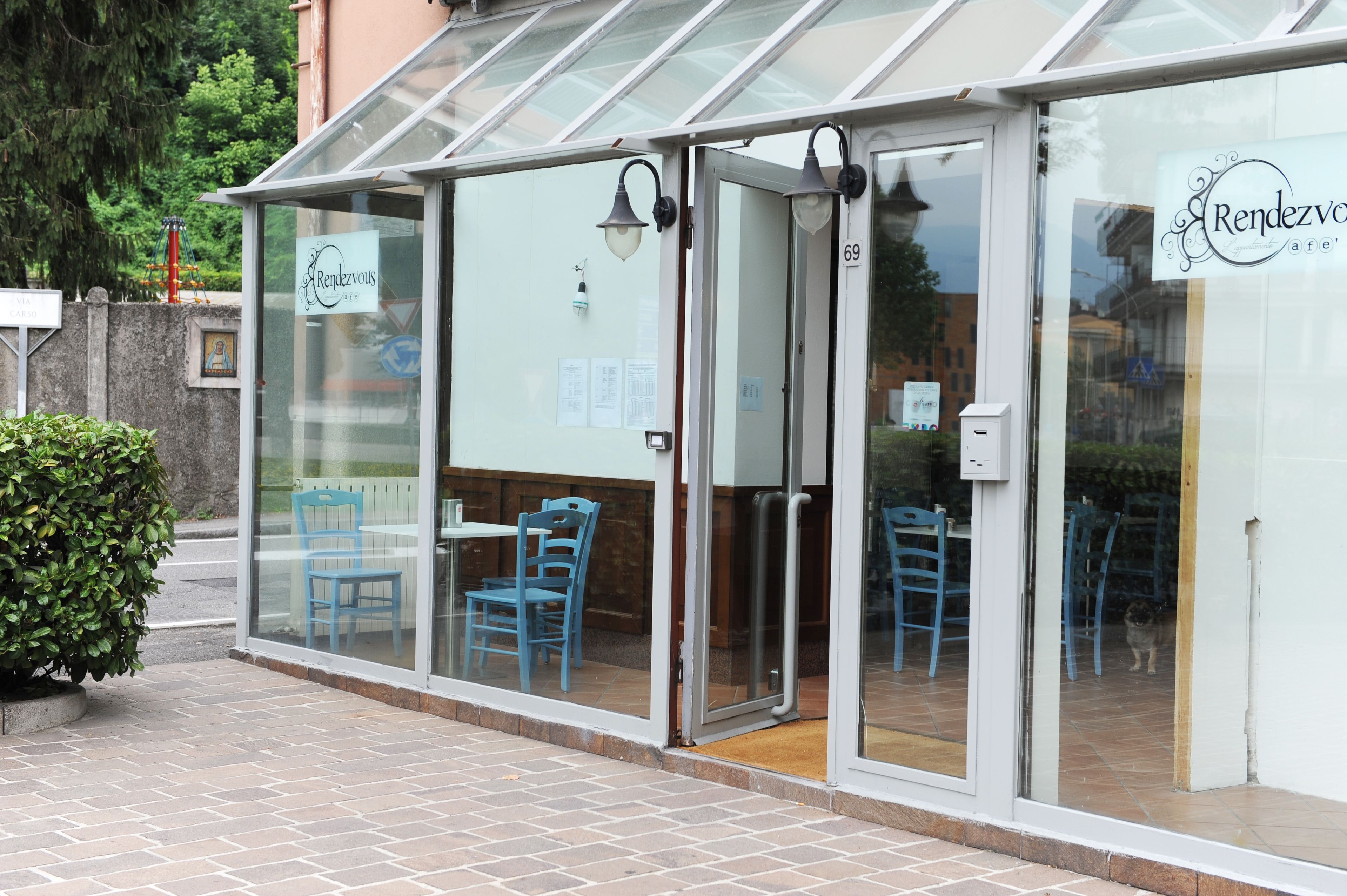 A glass-enclosed patio area with blue chairs and a table, a sign reading "Rendezvous cafe' 69", and a small dog visible through the glass doors.