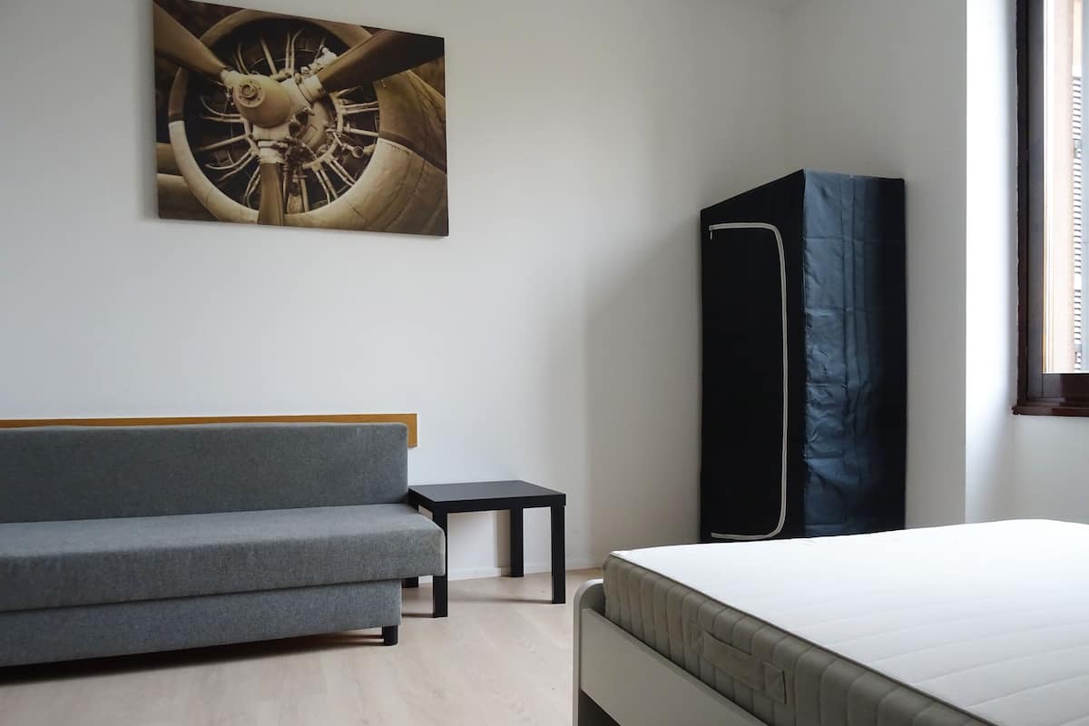 A modern bedroom with a grey sofa, a small black table, a bed with a white mattress, and a large painting of a mechanical wheel on the wall.