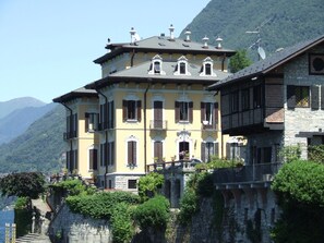 Un bâtiment historique doté de nombreux balcons et d’une façade en pierre, situé près d’un plan d’eau, avec des montagnes en arrière-plan.