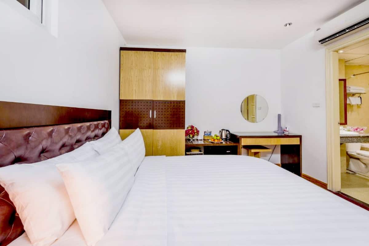 A hotel room with a large bed, a wooden wardrobe, a desk with a mirror, and a bathroom visible through an open door.