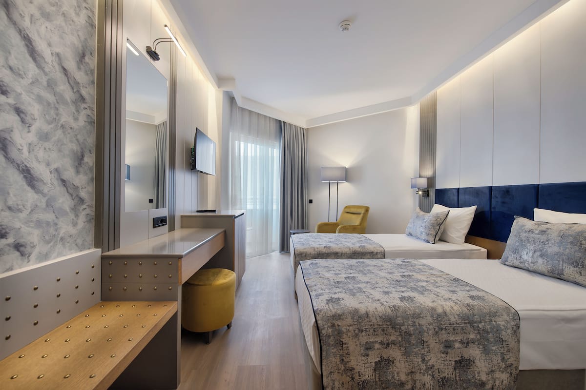 Une chambre d’hôtel moderne avec un mur en marbre, un banc en bois, deux lits avec des têtes de lit assorties, un bureau, une chaise et un grand miroir.