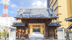 A traditional Japanese gate with a tiled roof and wooden pillars, leading to a building with a yellow facade and a sign with Japanese characters. - Sansuien (Kochi)