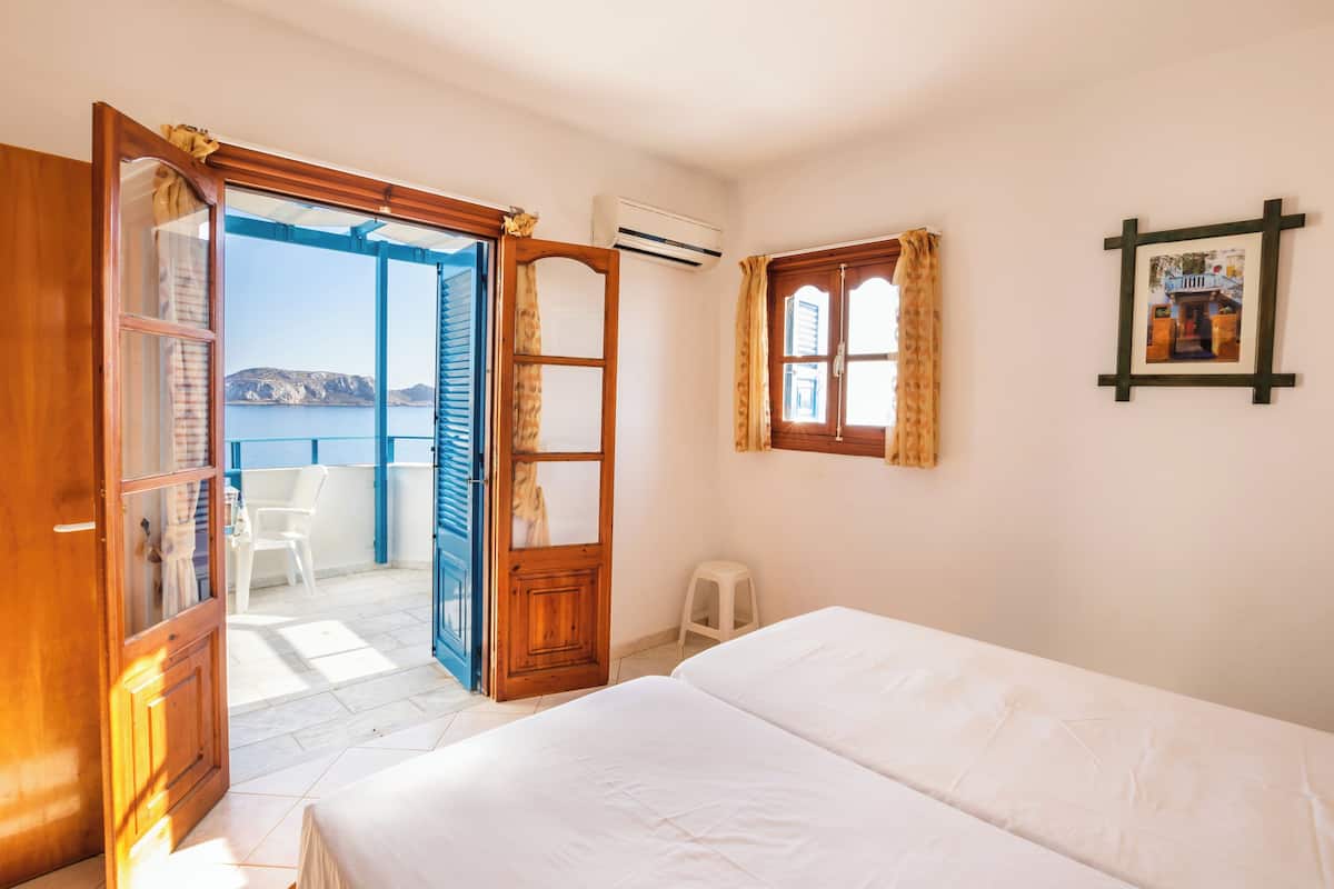 A bedroom with a bed, a window with curtains, a picture on the wall, and an open door leading to a balcony.