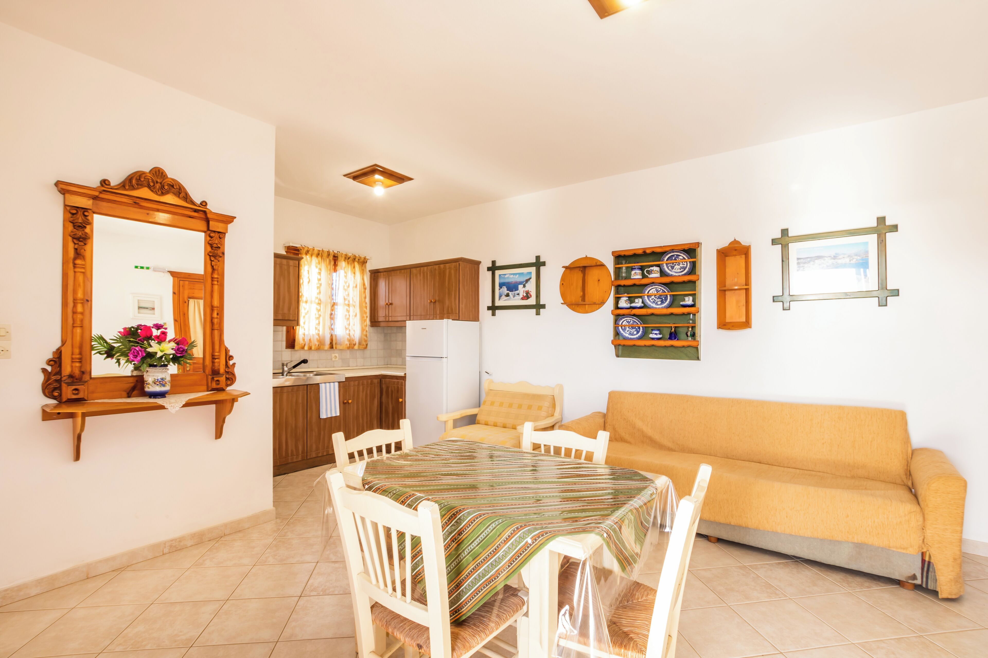 A dining area with a table and chairs, a sofa, a kitchen with cabinets and a refrigerator, and a mirror on the wall.