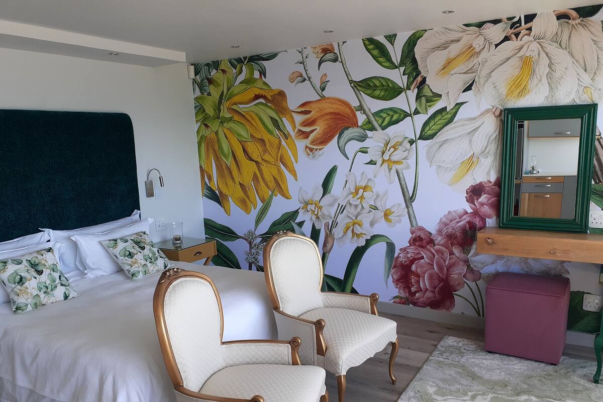 A bedroom with a bed, two chairs, a desk with a mirror, and a floral wall mural.