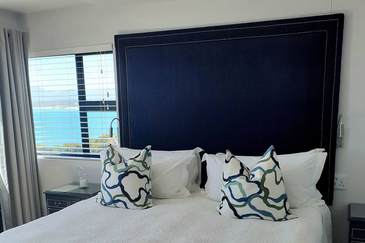 A large bed with a white textured bedspread and two decorative pillows, a dark headboard, and a view of the sea through a window with curtains.