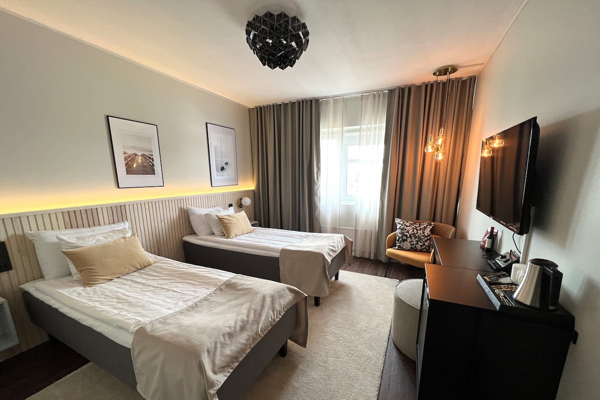Ein Hotelzimmer mit zwei Betten, einem Schreibtisch, einem Fernseher und einem Fenster mit Vorhängen.
