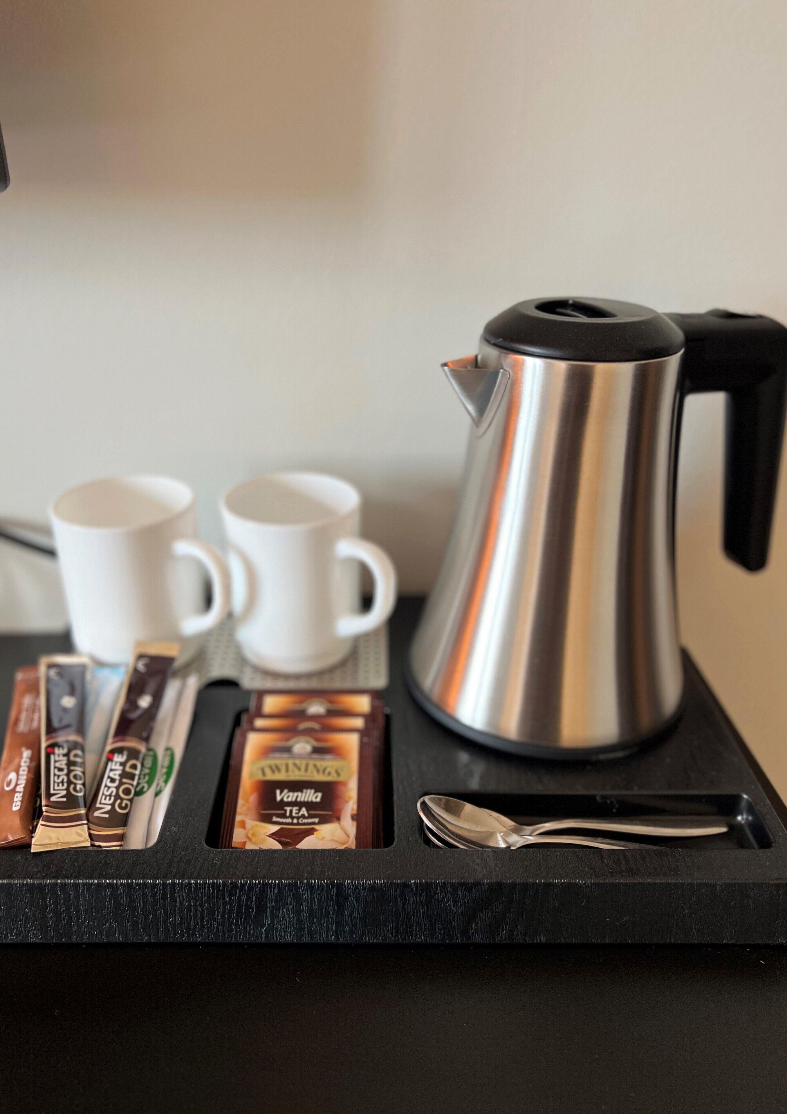 Una estación de café con una cafetera de acero inoxidable, dos tazas blancas, una cuchara y varios pods de café.