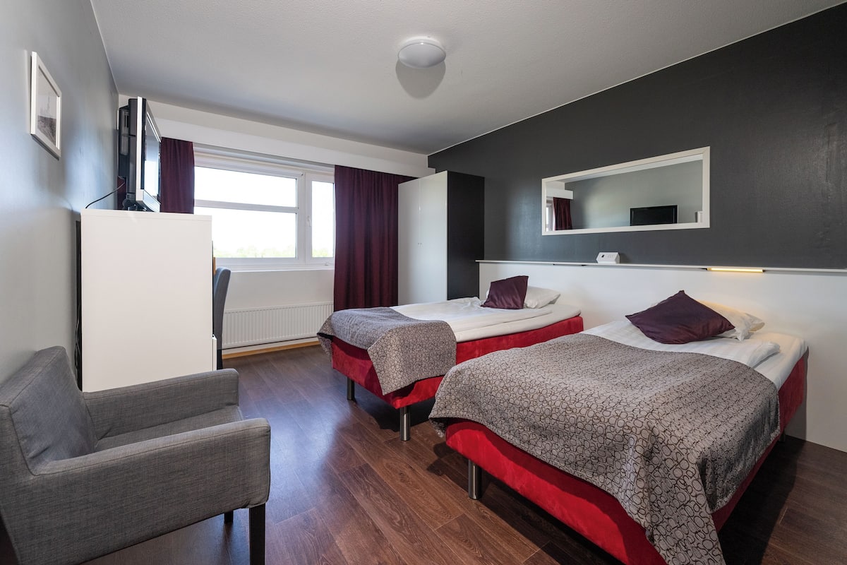 Ein Hotelzimmer mit zwei Betten, einem Sessel, einem Fernseher und einem Fenster mit Vorhängen.
