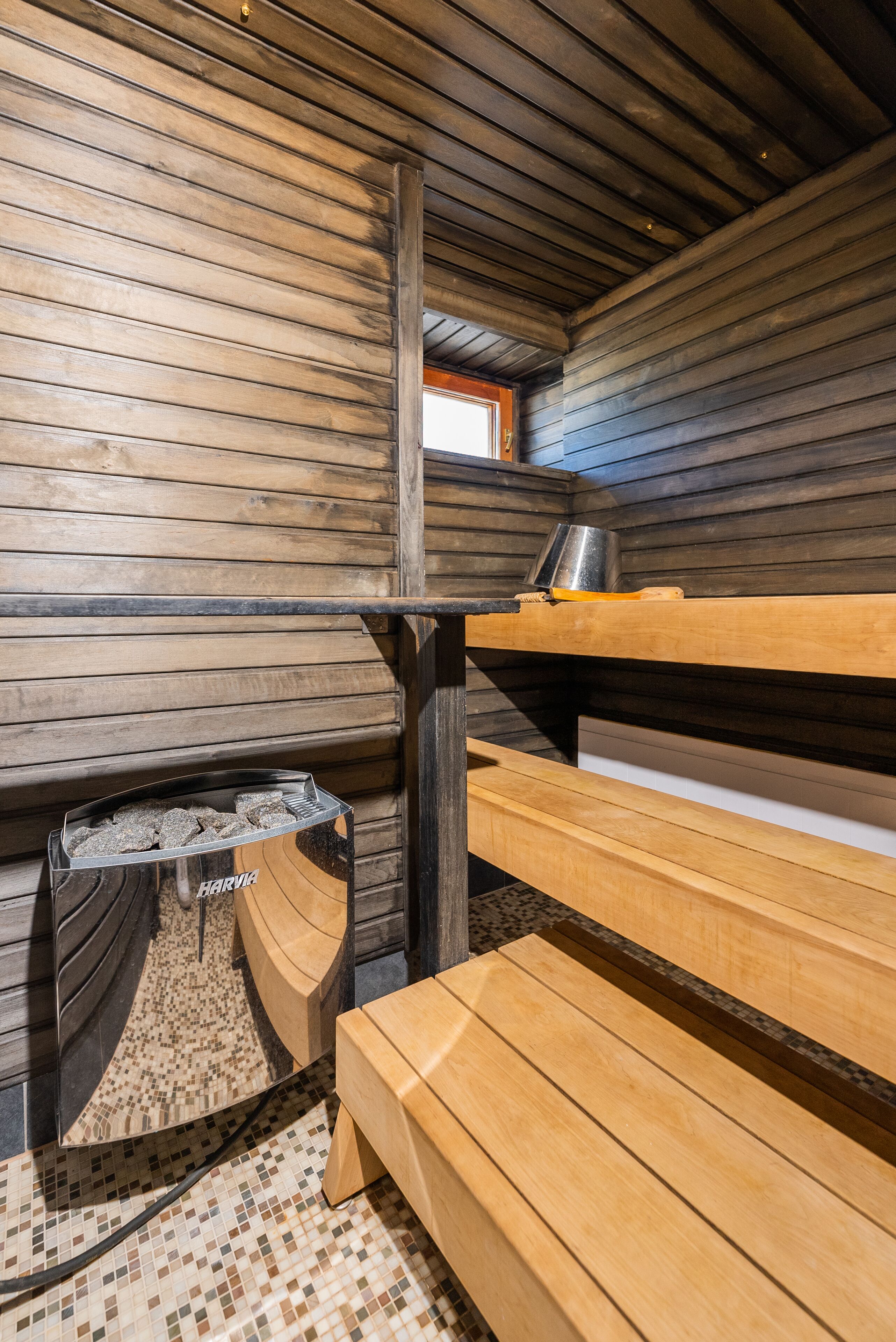 A sauna room with wooden benches, a bench with a metal container, and a tiled floor.