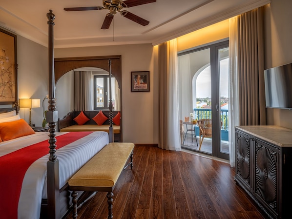 A hotel room with a four-poster bed, a bench, a balcony with wicker chairs, and a TV.
