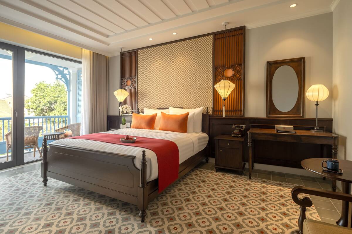 A hotel room with a bed, a desk, a chair, a small table, a mirror, and a balcony with outdoor furniture.