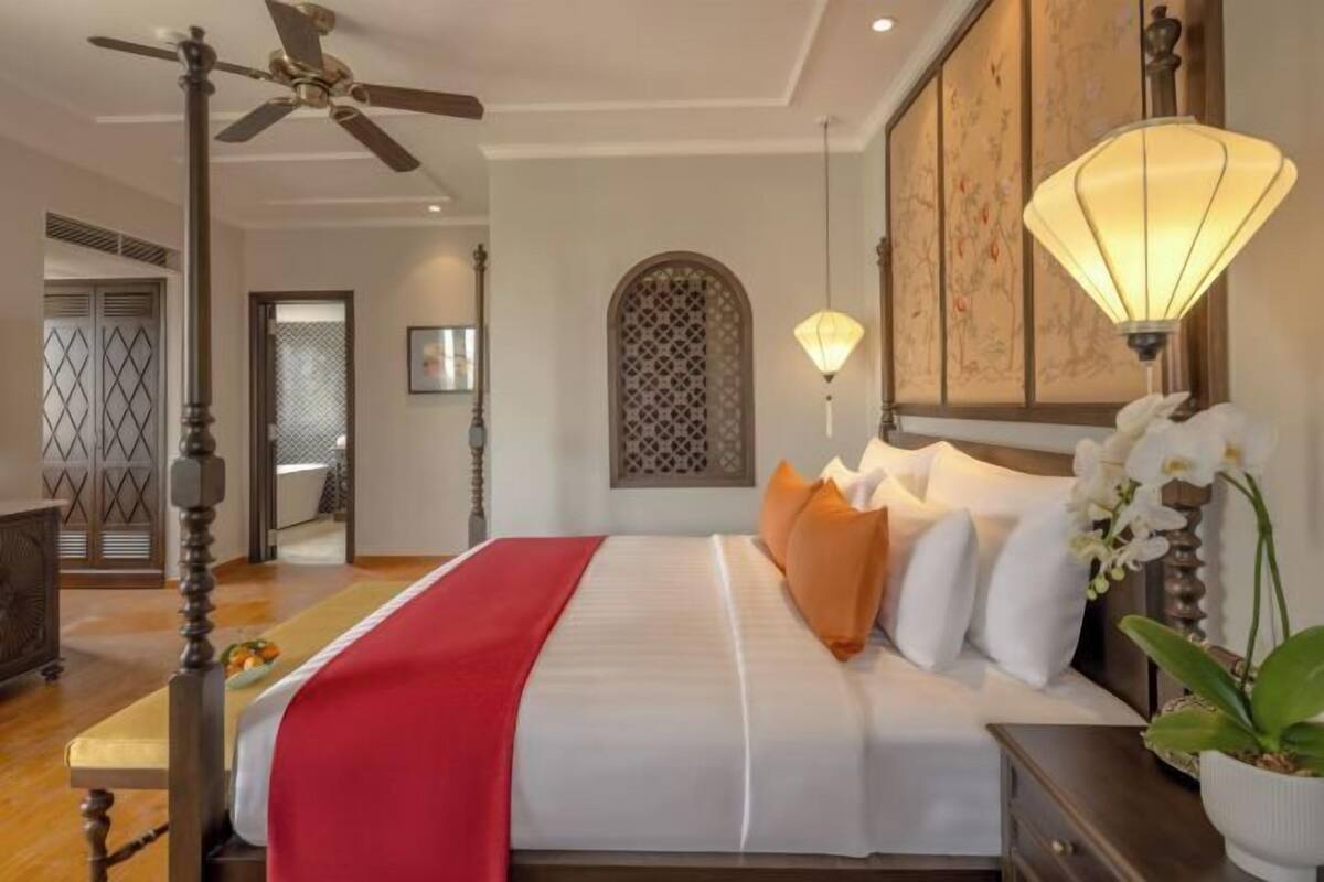 A hotel room with a bed, a ceiling fan, a nightstand, a lamp, and a decorative wall panel.