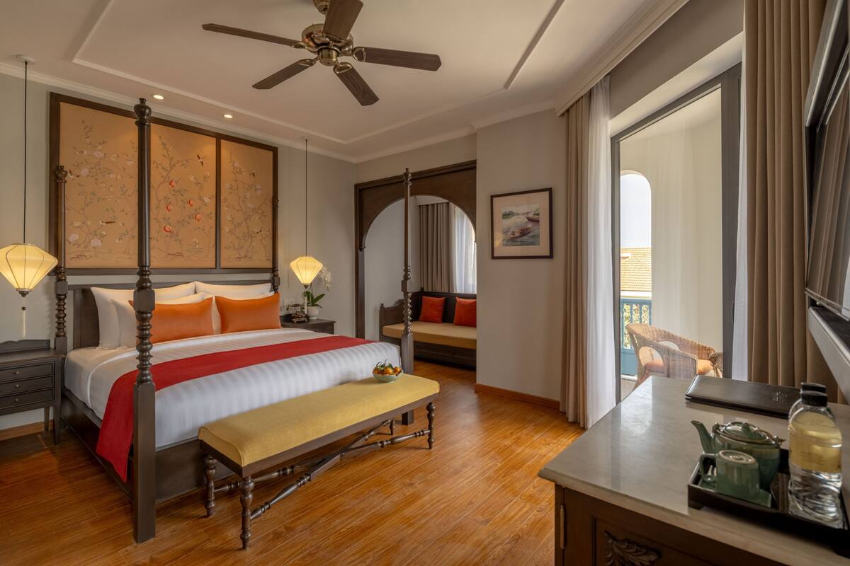 A hotel room with a four-poster bed, a desk with a teapot and bottle, a balcony view, and a traditional Japanese-style painting on the wall.
