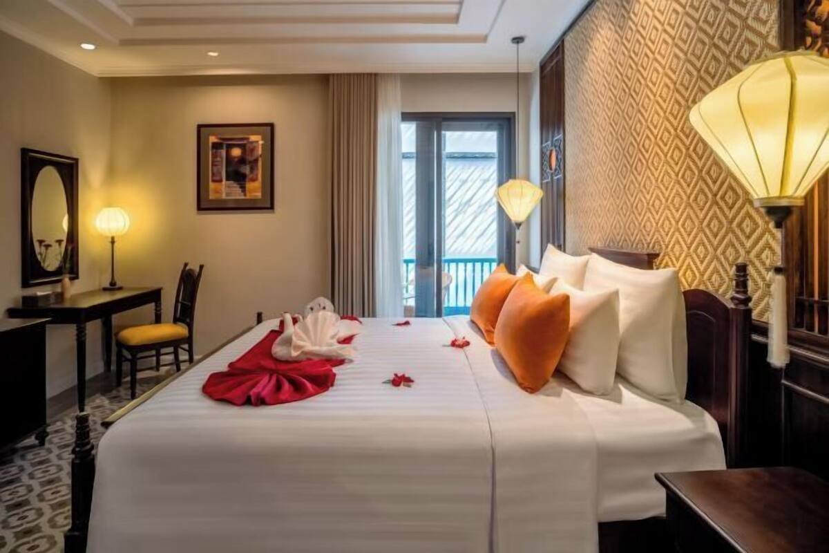 A hotel room with a bed, a desk, a chair, a lamp, and a view of the outside.
