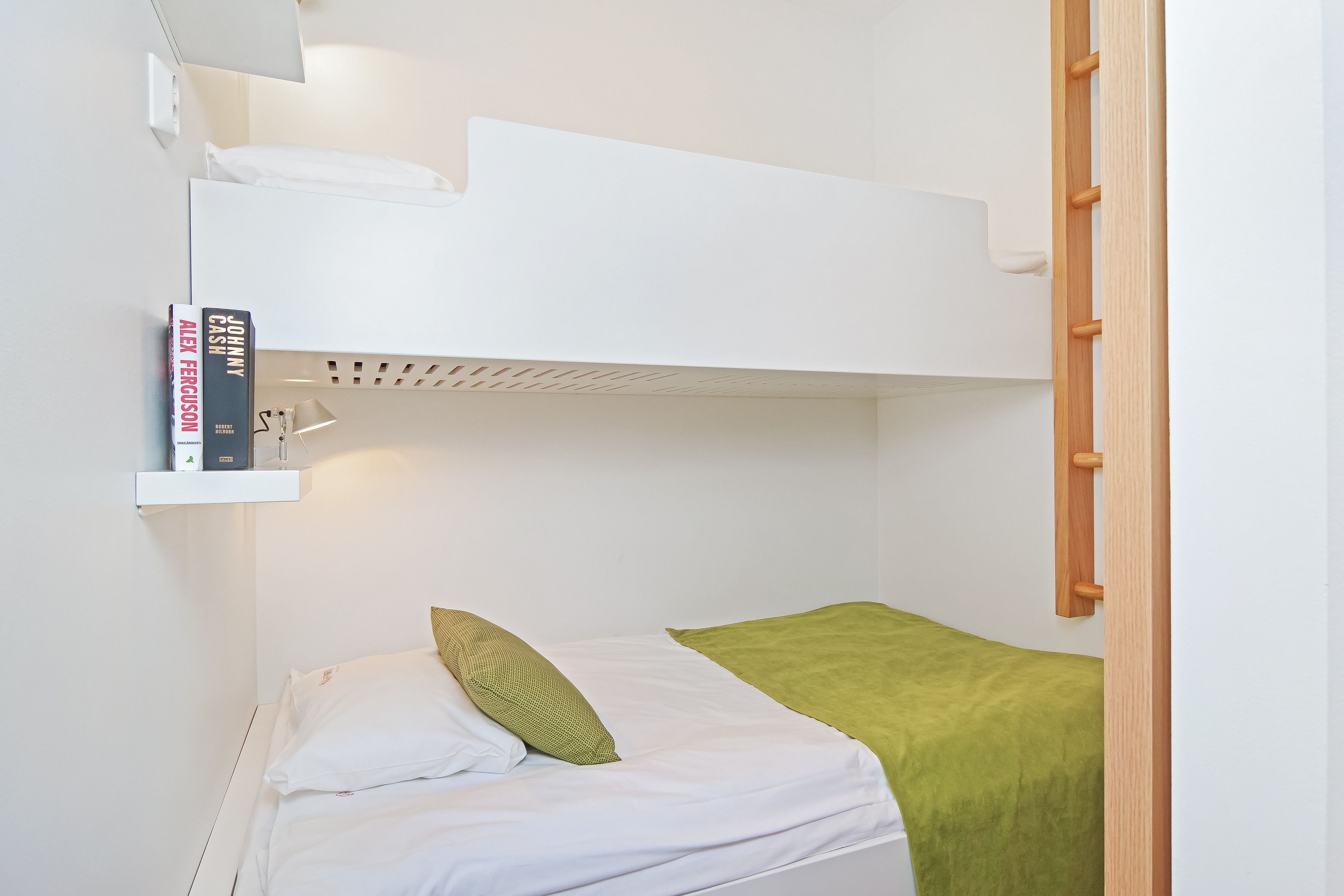 Una cama litera con un edredón verde, ropa de cama blanca y un estante con libros.