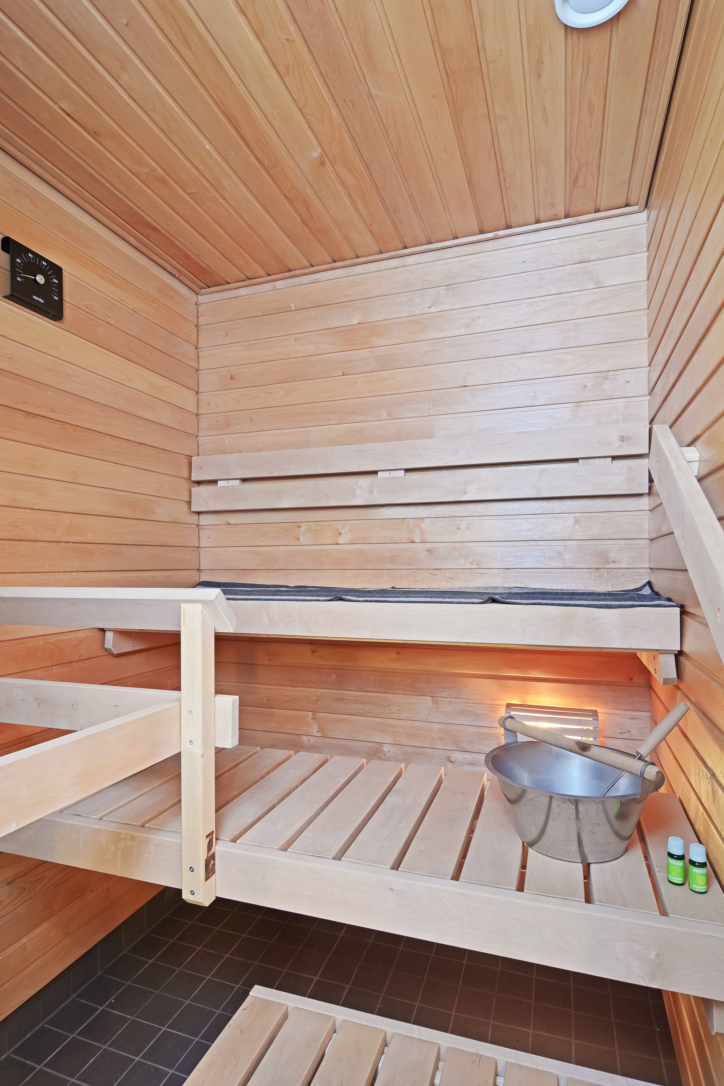 A wooden sauna with a bench, a bucket, and two bottles on the floor.