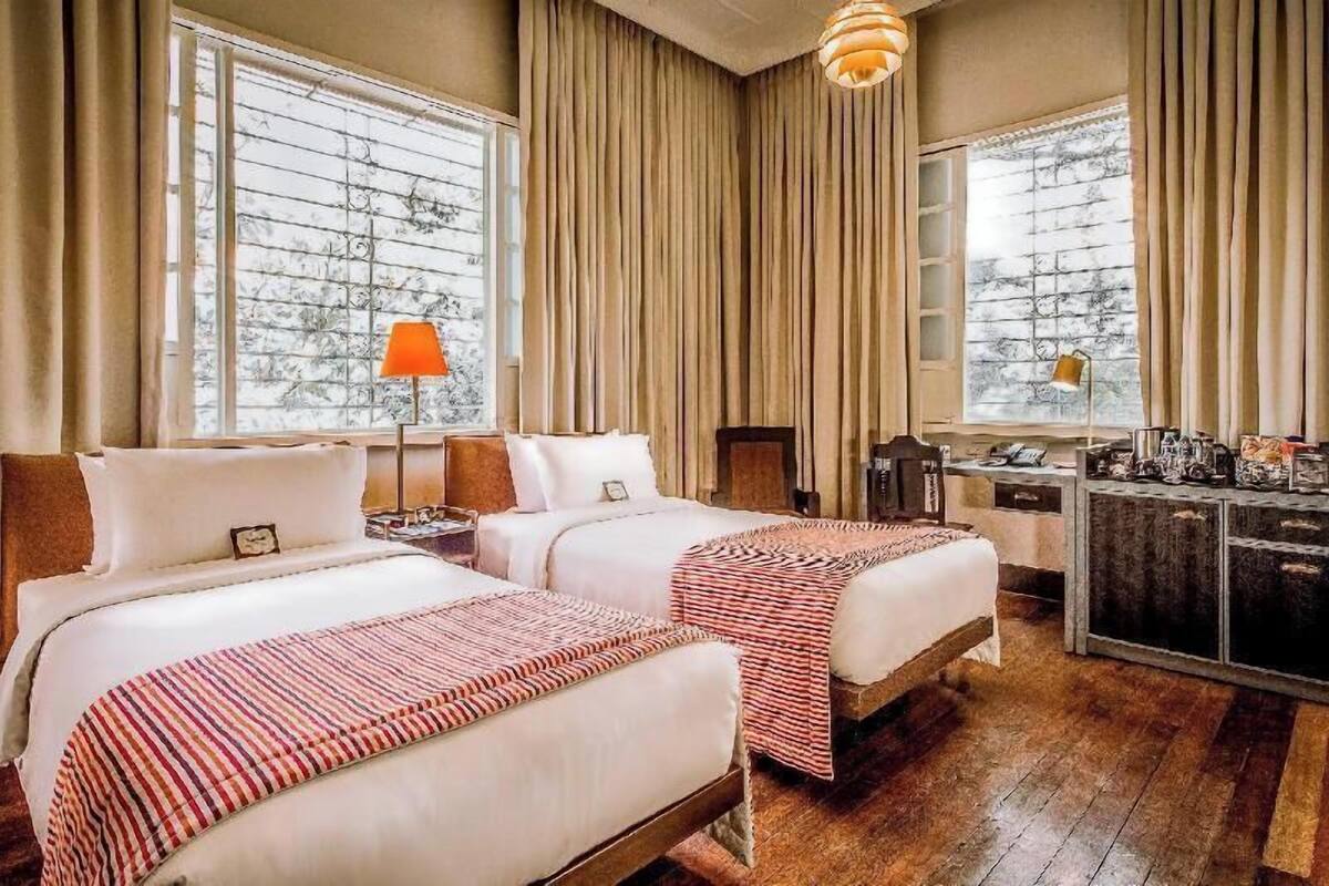 A hotel room with two beds, a desk, a chair, a lamp, and a view of trees through the window.