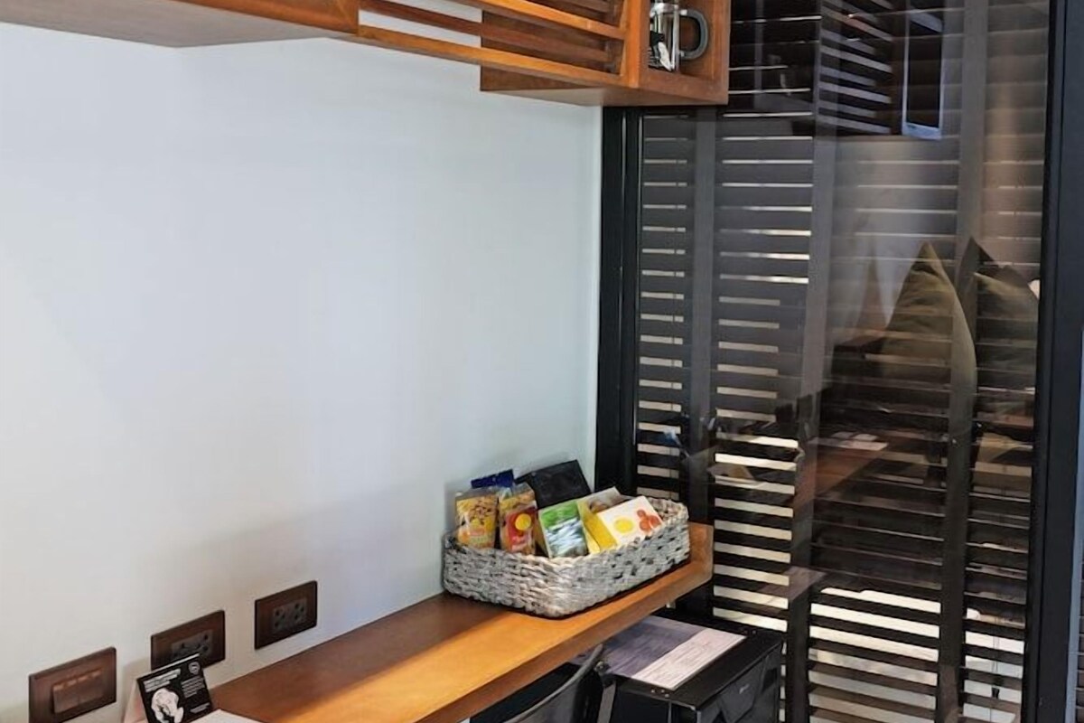 Habitación de hotel moderna con un escritorio de madera, una silla, un refrigerador pequeño y un separador de vidrio.
