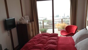Une chambre d’hôtel avec un lit, une télévision, un fauteuil rouge et une vue sur le paysage urbain, visible à travers de grandes fenêtres.