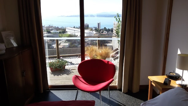 Une chambre d’hôtel avec un fauteuil rouge, un bureau en bois avec une lampe et une vue sur le paysage urbain et l’eau.