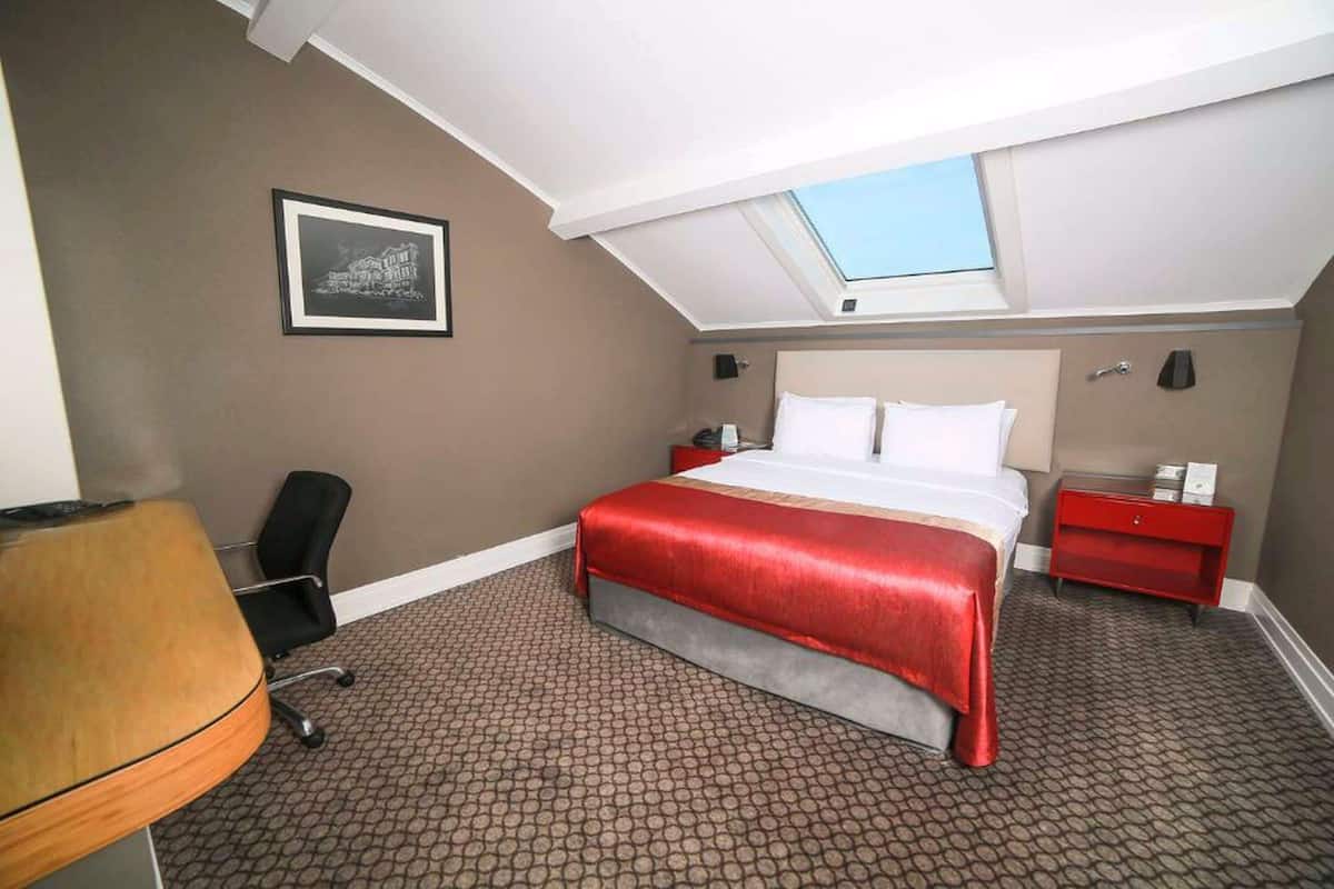 Une chambre d’hôtel avec un puits de lumière, un lit recouvert d’une courtepointe rouge, un bureau avec une chaise et un tableau encadré au mur.