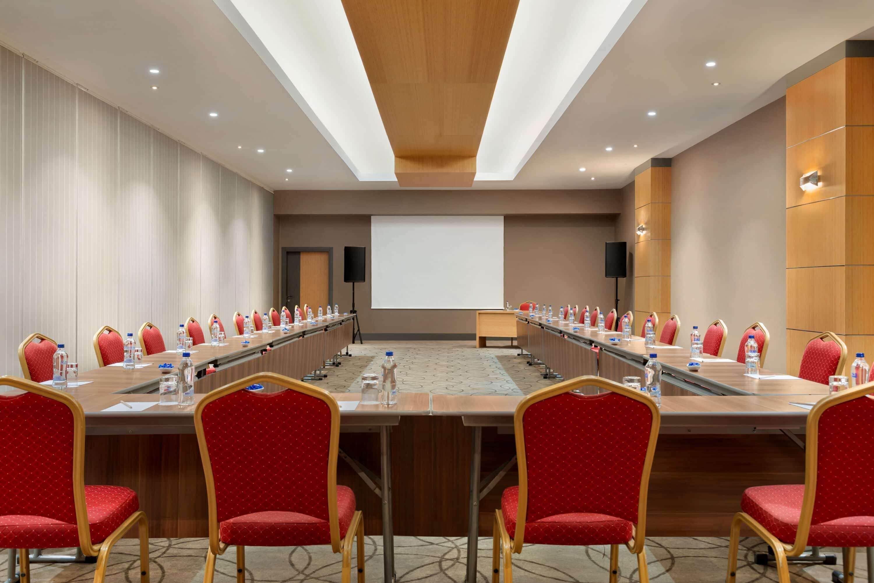Una sala de conferencias con una larga mesa, sillas rojas, botellas de agua y una pantalla de proyector.