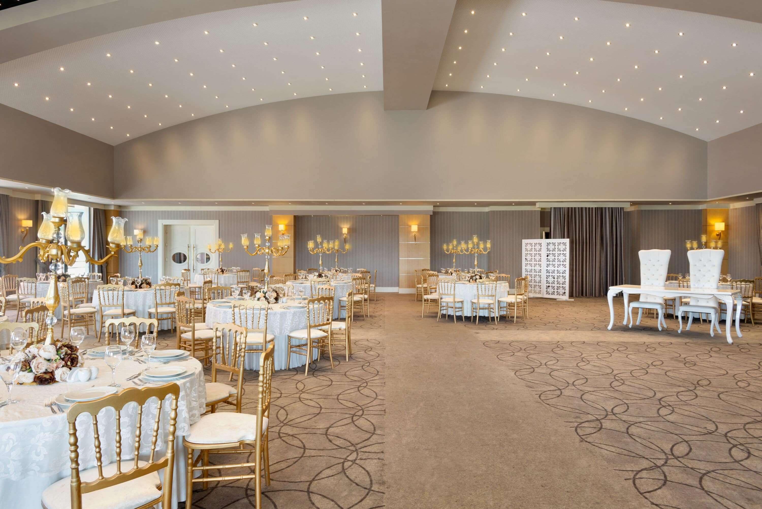 A spacious banquet hall with multiple round tables, gold chairs, and elegant chandeliers.
